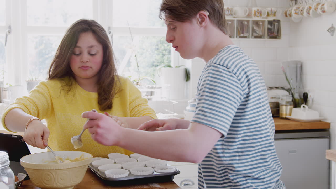 젊은 다운 증후군 부부가 집에서 주방에서 종이 컵케이크 케이스에 혼합물을 넣고 있습니다.