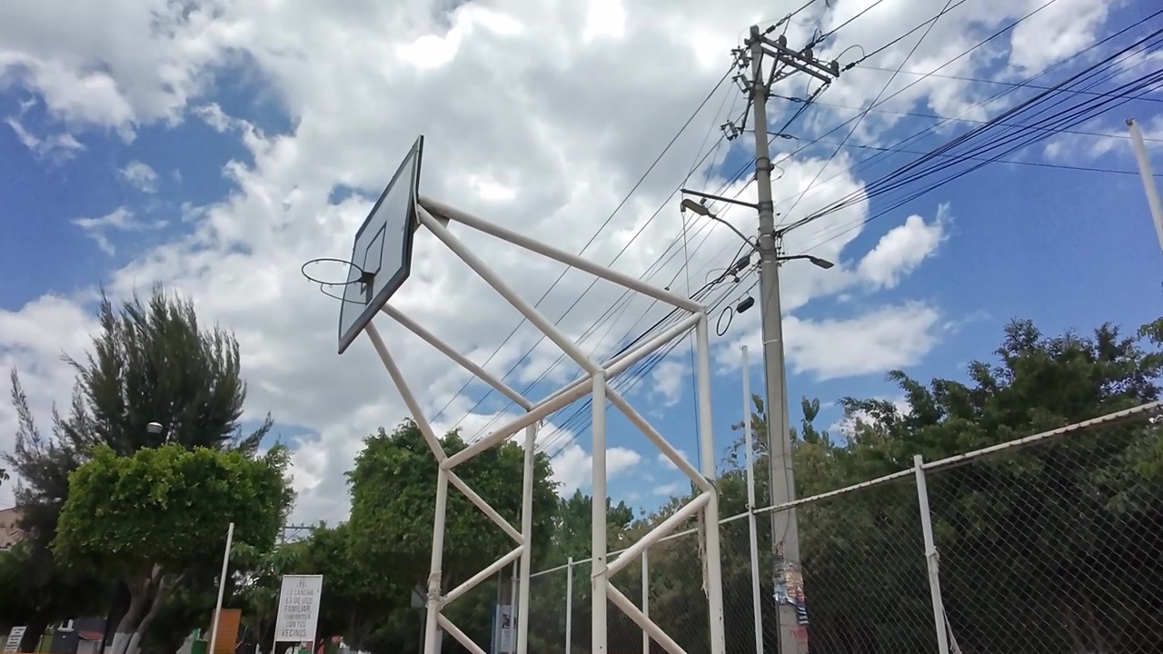 Cesto de basquete ao lado de uma cerca de arame e poste de eletricidade num campo exterior em Huatulco, com árvores e linhas de energia visíveis sob um céu azul parcialmente nublado.