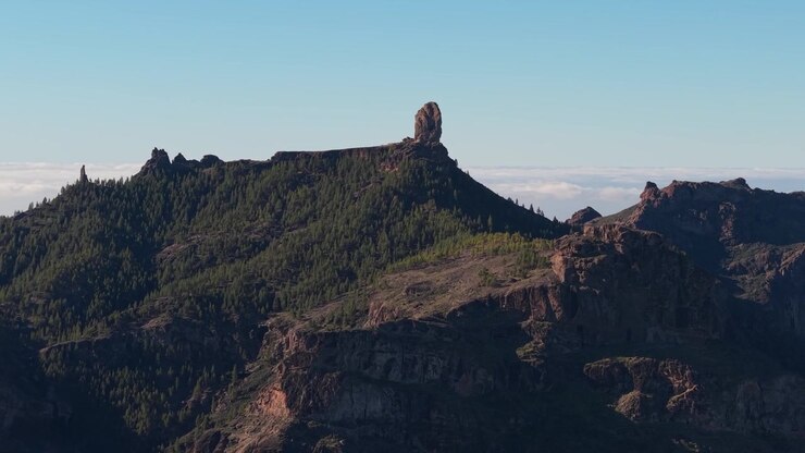 Luftfoto af Roque Nublo, den ikoniske vulkanske monolit på Gran Canaria, der står på et fyrretræsbevokset plateau over et hav af skyer under en klar blå himmel