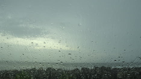 Closeup-raindrops-water-droplets-trickling-down-on-wet-clear-car-window-glass-during-heavy-rain-against-beach-view-in-rainy-day-monsoon-season