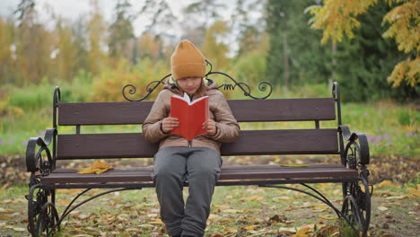 little girl wearing mustard beanie reading red cover book on park bench under golden foliage autumn leaves beside her as she flips pages glancing at surroundings