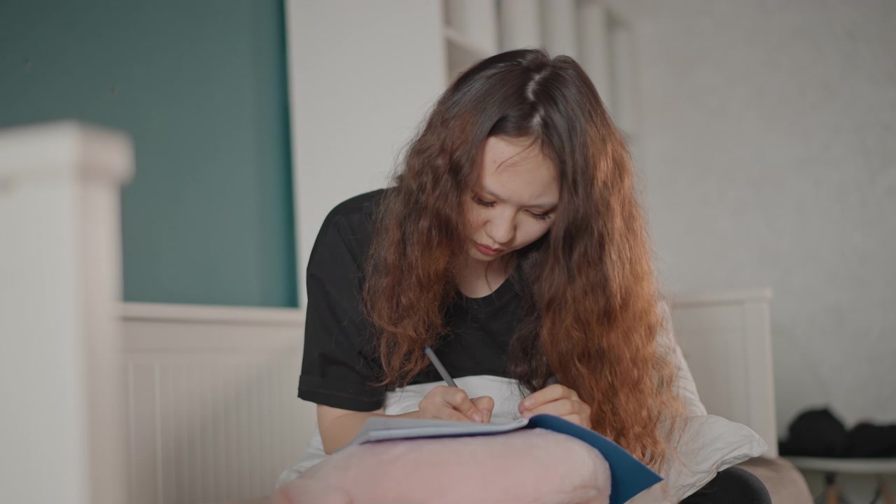 Giovane donna seduta sul letto si china in avanti mentre scrive su un quaderno, la penna si muove sulla pagina con intensa concentrazione, in un ambiente tranquillo della camera da letto, con luce interna soffusa e un'atmosfera di routine domestica rilassata.