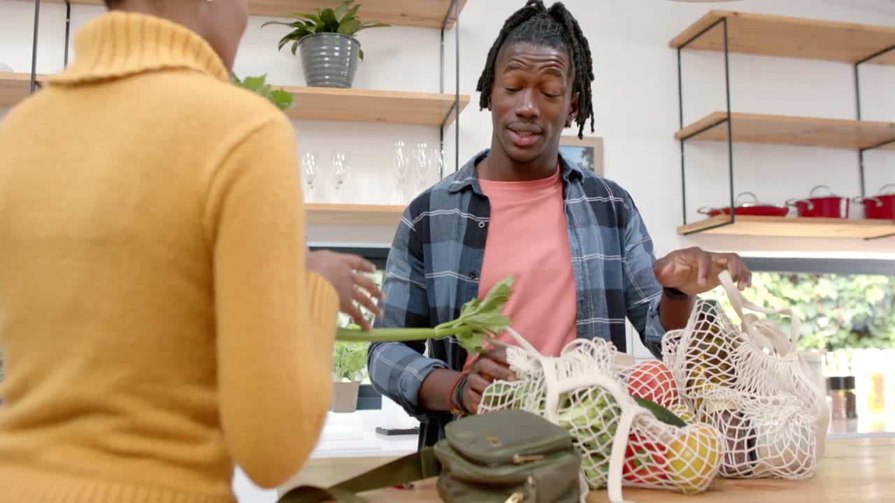 feliz pareja afroamericana desempaquetando comestibles en la cocina, cámara lenta