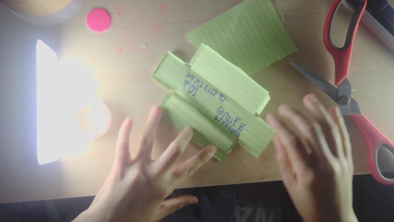 Una ripresa dall'alto mostra le mani di un bambino preadolescente che usano forbici rosse per tagliare carta a righe verde piegata, mentre realizzano un lavoretto fai-da-te di squishy di carta su una scrivania di legno illuminata da una brillante lampada a LED.