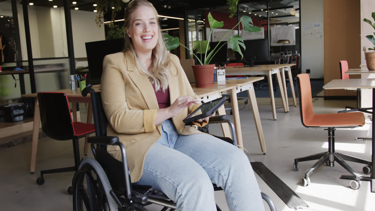 retrato de una feliz mujer de negocios caucásica en silla de ruedas usando una computadora portátil en la oficina, en cámara lenta