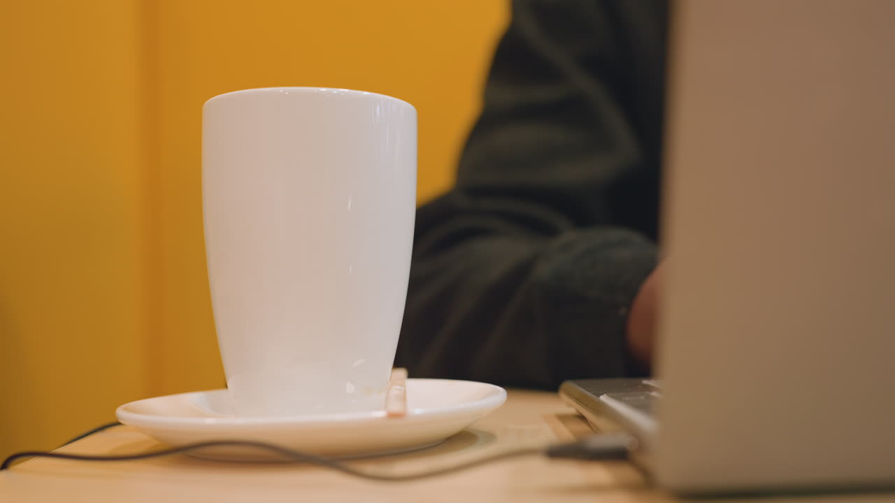 鮮やかな黄色のカフェで、白いカップの隣で黒い手がノートパソコンでコーディングしている。キーボードのクリック音から締め切りのプレッシャーが伝わり、USBケーブルとマウスが見える。集中したエネルギーとカフェインによる覚醒が感じられるモダンなワークスペース。