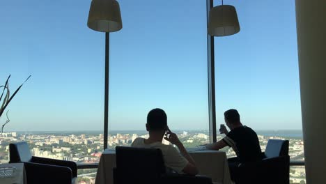 two men relaxing at a high-rise restaurant with city view