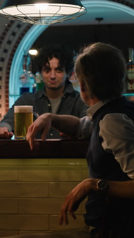 vertical shot of young bartender pours craft beer into glass and gives it to mature man. male friends clink glasses and drink beer sitting at the bar counter in stylish pub at night. concept of lifestyle and leisure.