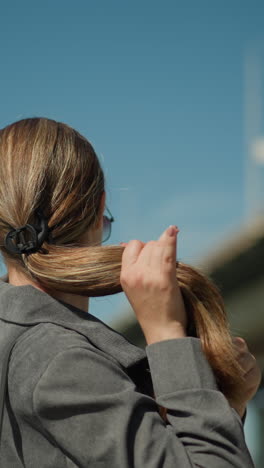 la dama se endereza el cabello mientras mira lejos cerca de un puente, ajusta la parte delantera de su cabello mientras se exhibe la longitud, con postes eléctricos borrosos y estructuras en el fondo