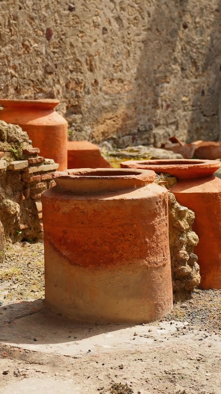 Primo piano verticale di giare di terracotta per la conservazione tra le rovine di Pompei, che mostra vasi di argilla, ghiaia illuminata dal sole, ruvide mura antiche e manufatti domestici romani conservati