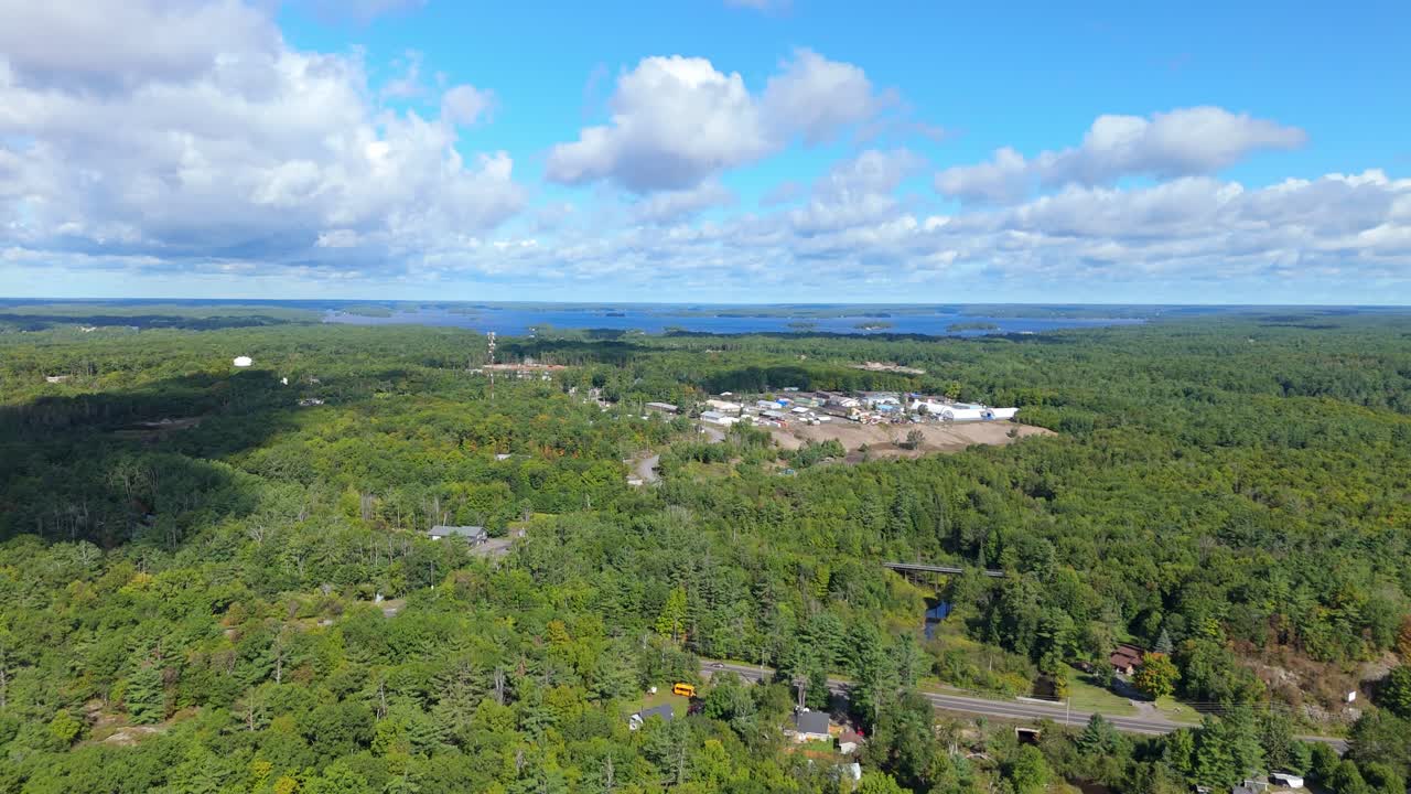 Aerial drone view of industrial buildings surrounded by dense forest with a lake visible in the distance near Gravenhurst, Muskoka, Ontario, showing rural industry within natural landscape