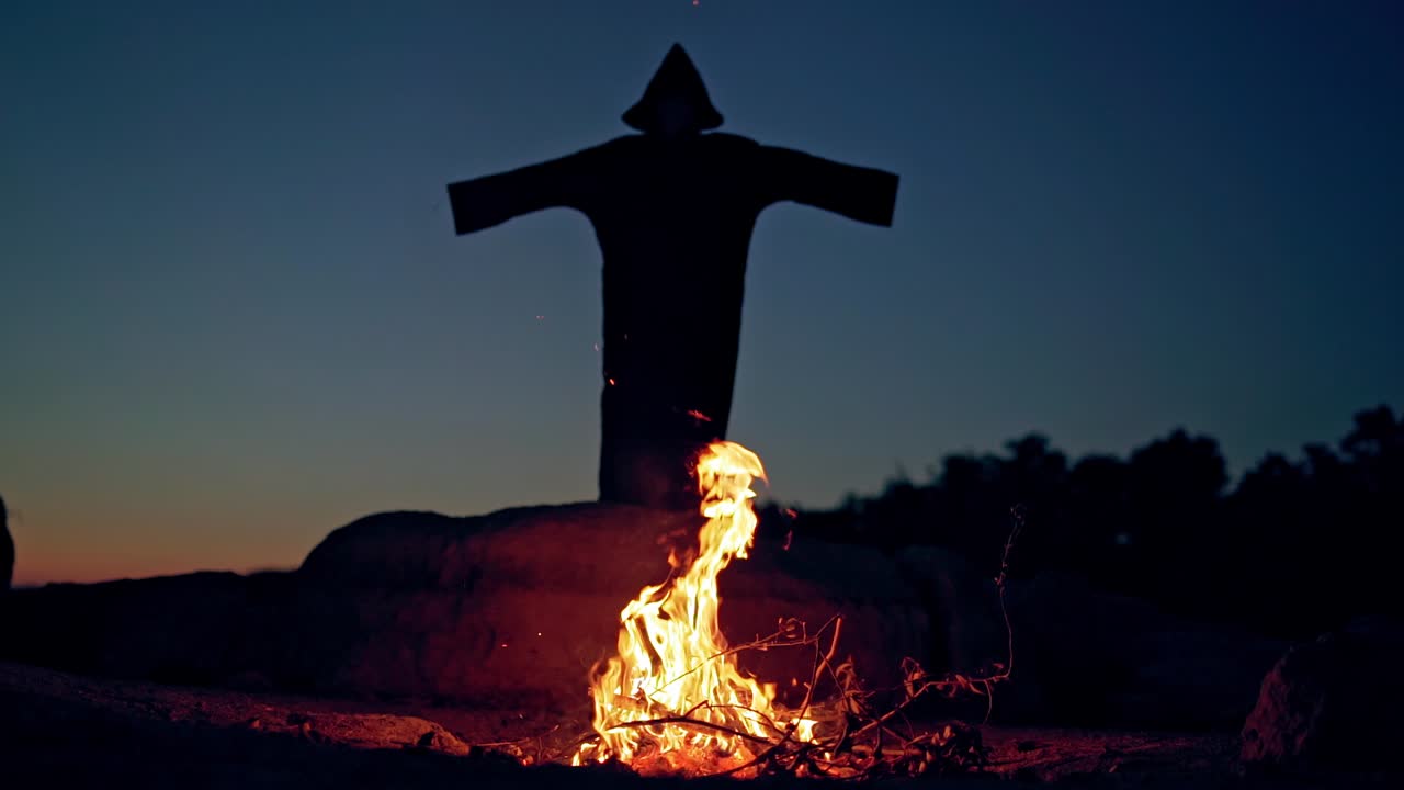Witch raising hands up. Black witch in black mantle is standing near fire