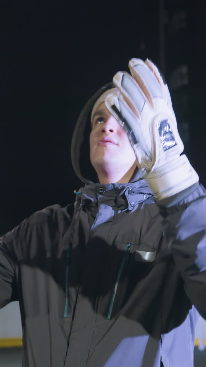 장갑을 입은 코치가 축구공을 공중으로 던지고 도시 경기장, 조명된 배경에서 야간 훈련 기간 동안 다시 잡습니다.