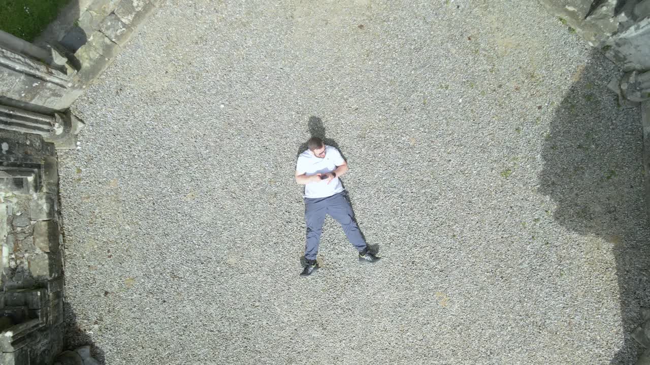 Lying on gravel in an ancient monastery, a man is immersed in his phone in Mellifont Abbey, Ireland