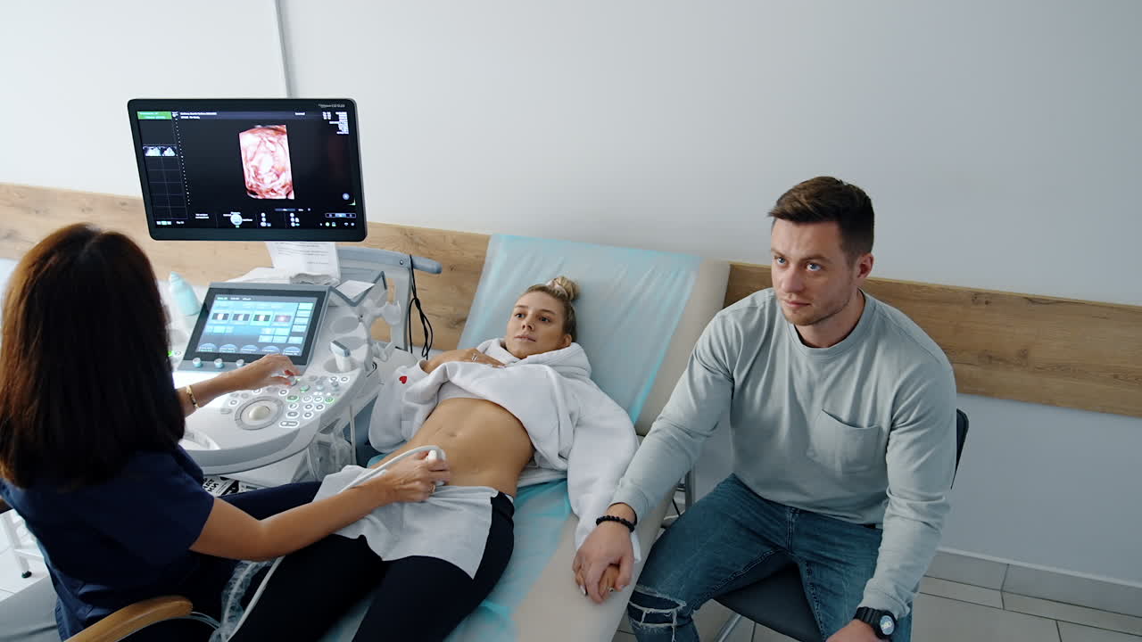 Expecting couple undergoing ultrasound check up in the hospital. Happy future parents looking focused on the screen. Brunette female doctor performs examination. Top view.