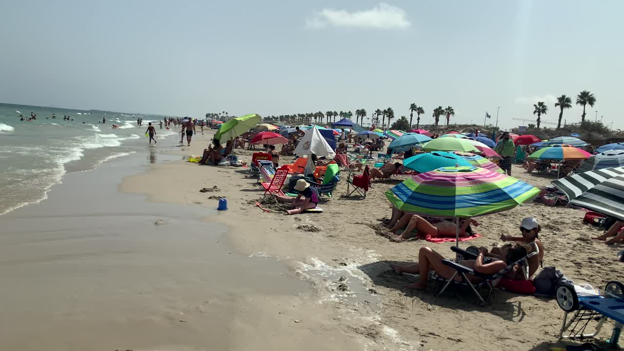 An array of colorful sun umbrellas dot the crowded beach, as people relax and enjoy the cool ocean breeze, soaking in the refreshing seaside atmosphere.