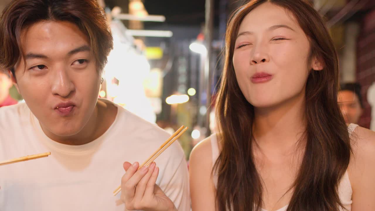 Smiling Asian couple eats noodles together at night market, warm lighting, handheld camera, candid mood