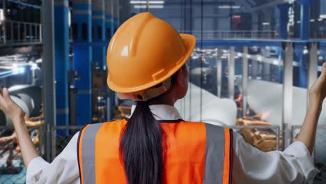 vista posterior cercana de una ingeniera asiática con casco de seguridad extendiendo los brazos mientras está de pie en una fábrica de fabricación de turbinas eólicas