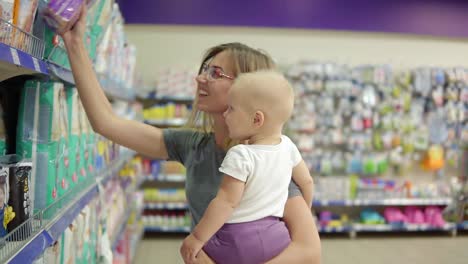 smiling mother in a section for children in the supermarket holding her child in her arms while choosing diapers on the shelves