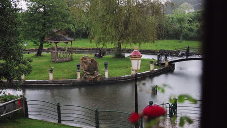 Scenic-garden-with-a-wooden-gazebo,-decorative-bridge-over-a-pond,-and-lush-greenery-on-a-rainy-day,-capturing-a-peaceful-outdoor-ambiance