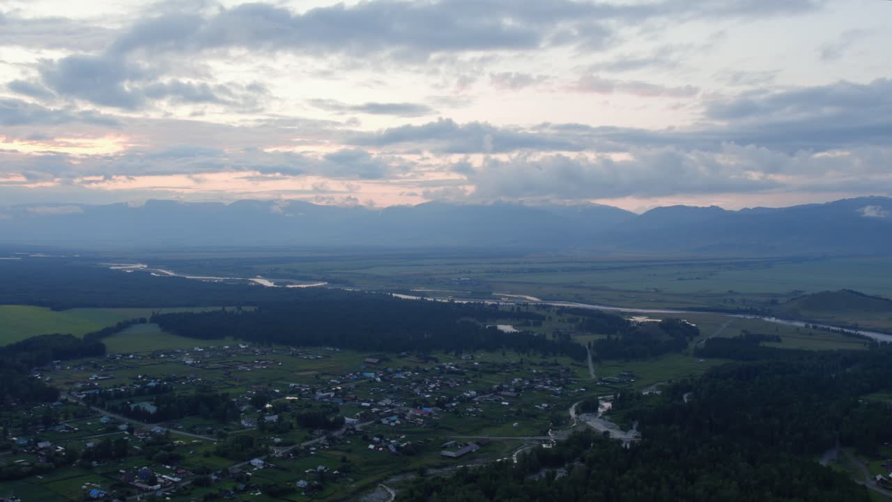 ロシア、アルタイ共和国のザムルタ村とカトゥン川、そして劇的な夕暮れの雲の下に広がる山間の谷のドローン映像。風光明媚な田園風景。