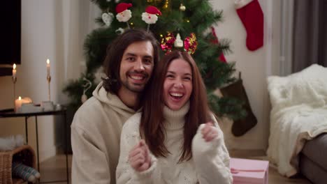 retrato de un feliz chico moreno que se sienta con su novia morena en un suéter blanco que mueve sus manos hacia adelante y hacia atrás al ritmo de la música cerca del árbol de año nuevo en una habitación decorada para el año nuevo en invierno