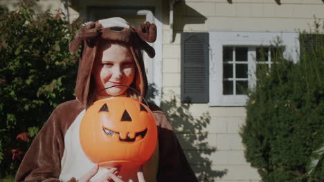 retrato de una niña en un disfraz de ciervo con una canasta de dulces en forma de calabaza. de pie frente a su casa, lista para ir por dulces