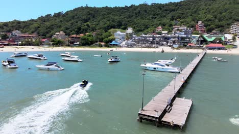 escena aérea de aviones no tripulados de bajo vuelo sobrevolando lanchas rápidas, yates y barcos de lujo con muelle en playa paradisiaca en canajure florianópolis santa catarina