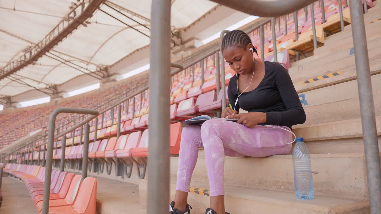 black student studying on stadium bleachers, writing notes in notebook, wearing pink leggings and earphones, water bottle beside, empty rows and warm sunlight creating calm focused atmosphere, candid