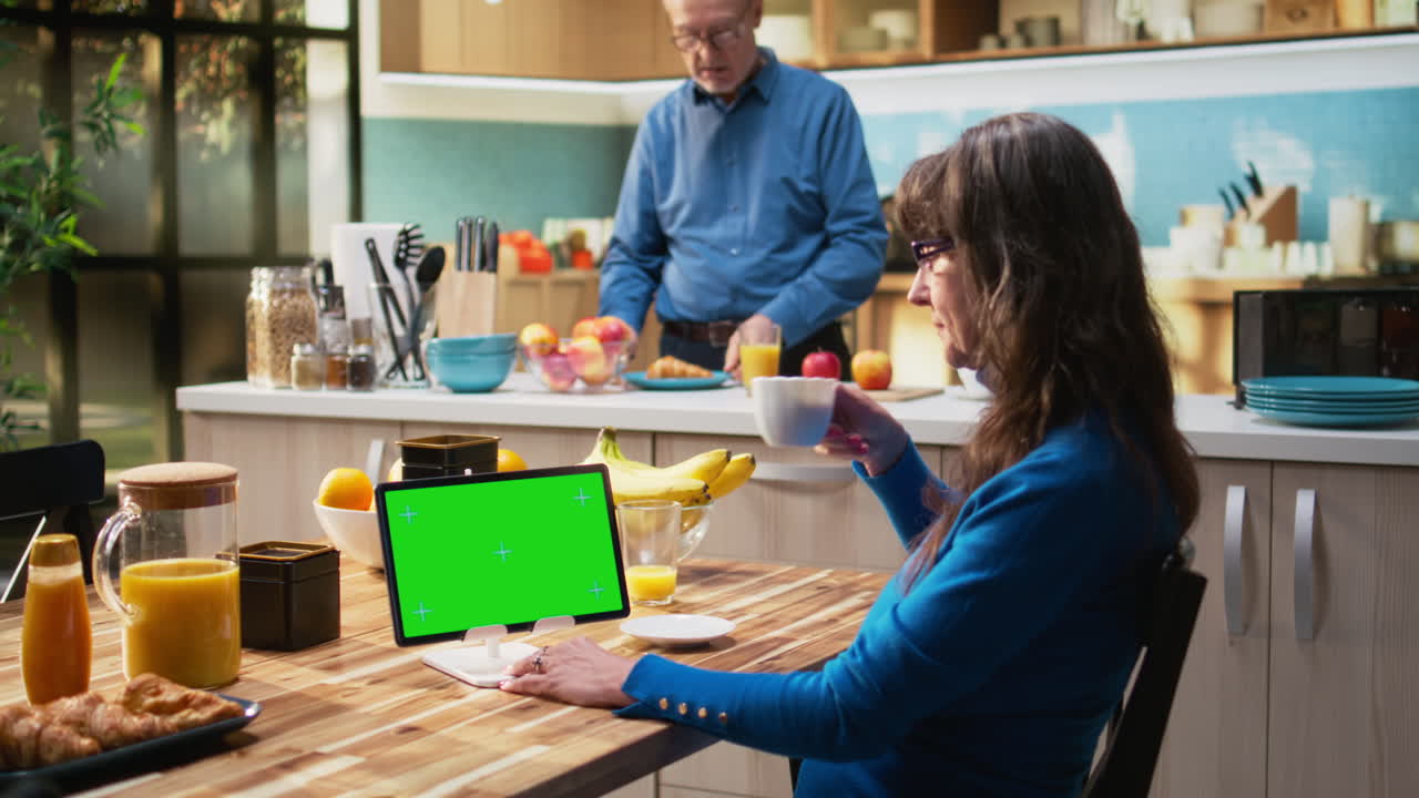 Green screen tablet with senior woman sitting at kitchen table with coffee cups