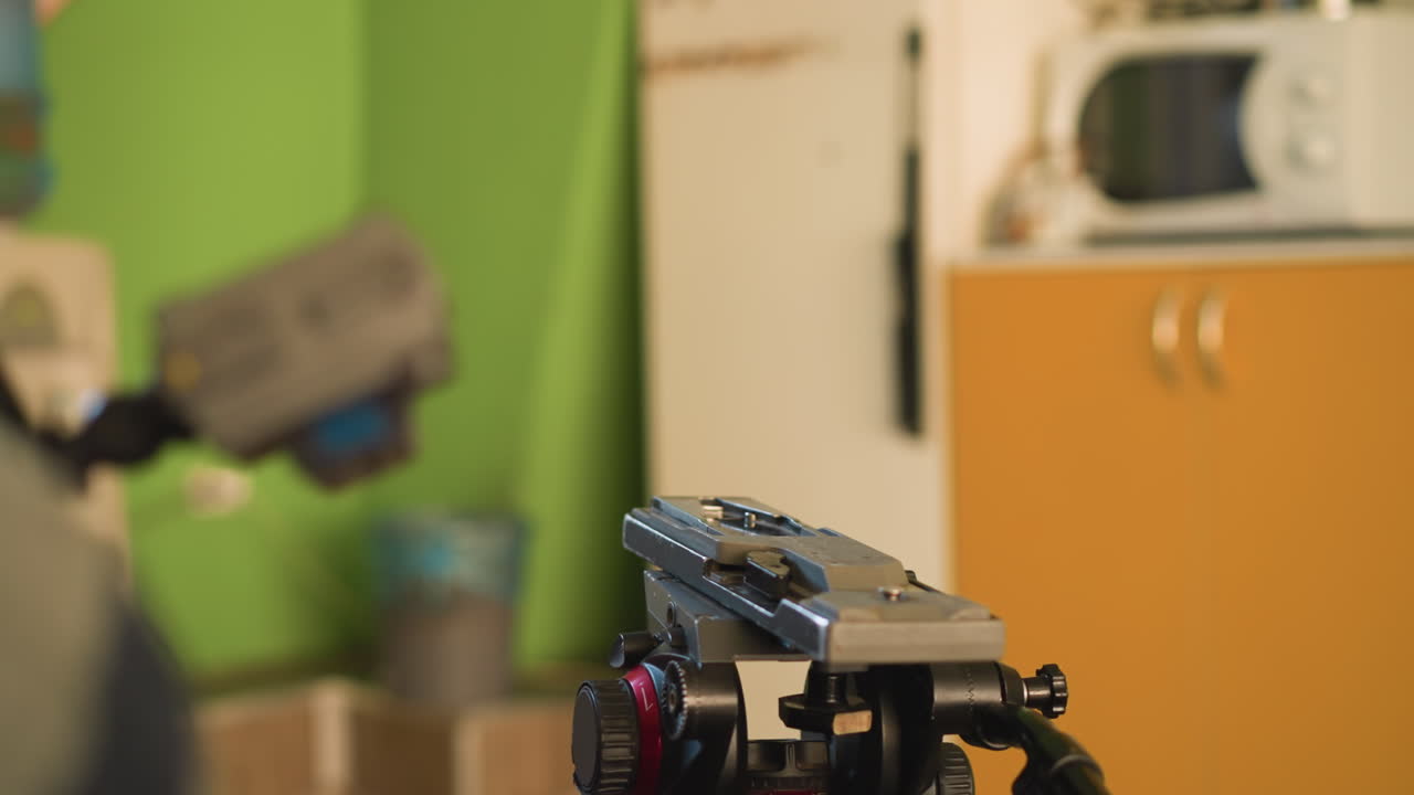 Close-up of hand adjusting camera lens on tripod mount. Background features water dispenser, orange cabinet, and blurred elements. Camera setup for filming in a home or studio setting