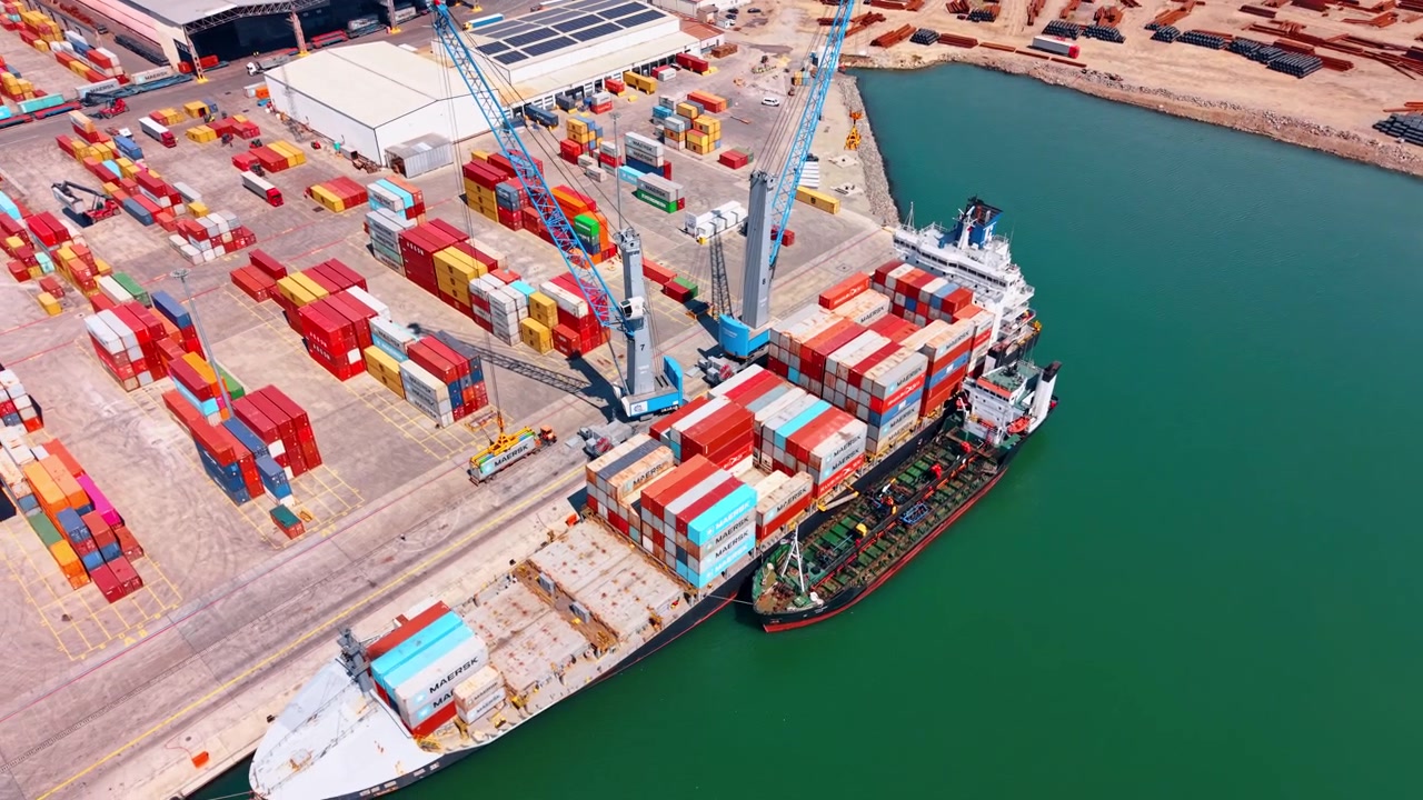 Burgas, Bulgaria, 1 May 2026: Cargo vessel and service ship berthed at container terminal. Aerial view of two vessels along quay, cranes, container stacks, warehouse roofs and green harbor water.