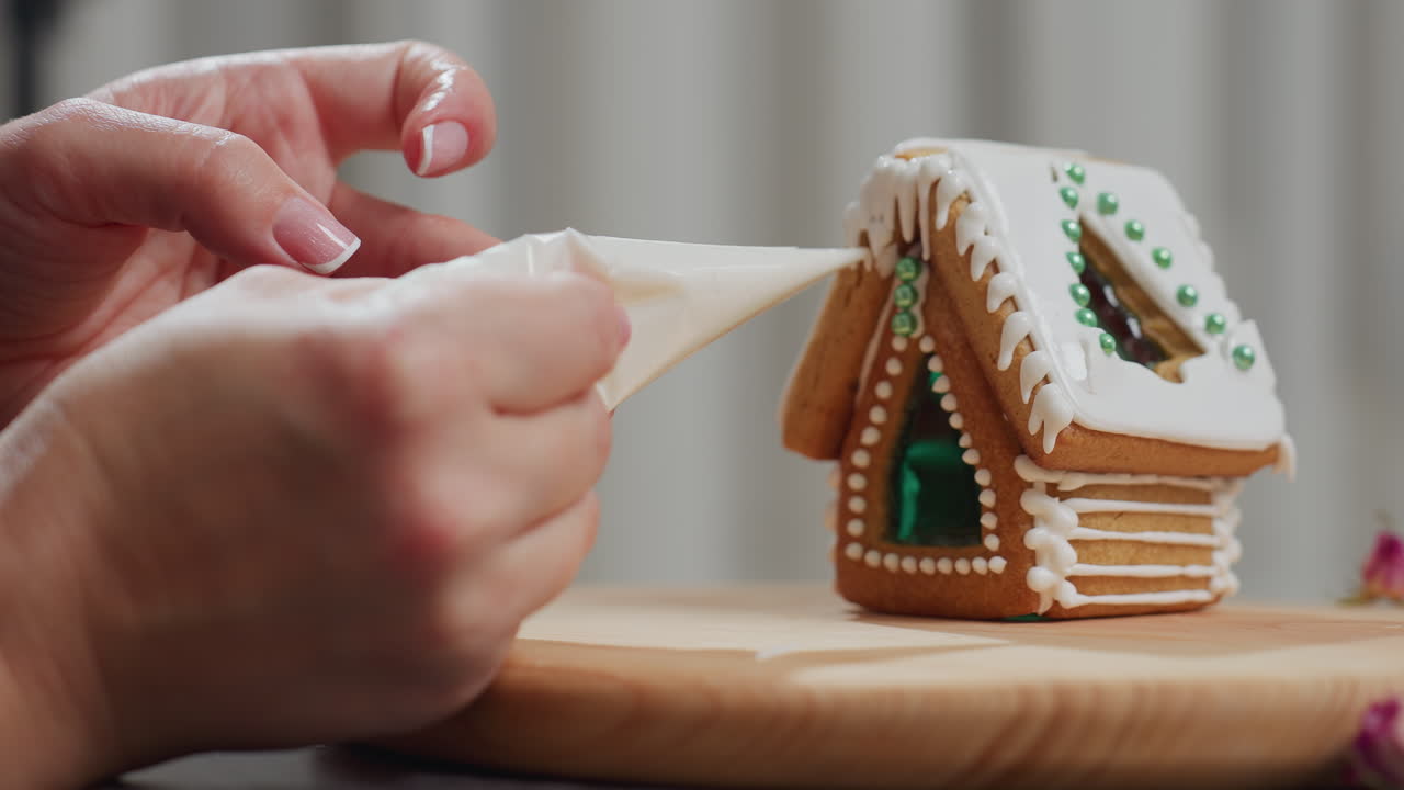 primer plano del proveedor decorando la casa de pan de jengibre recién horneado con detalles intrincados de glaseado, decoraciones de cuentas agregan encanto festivo mientras las manos aplican cuidadosamente el glaseado en la bandeja de madera