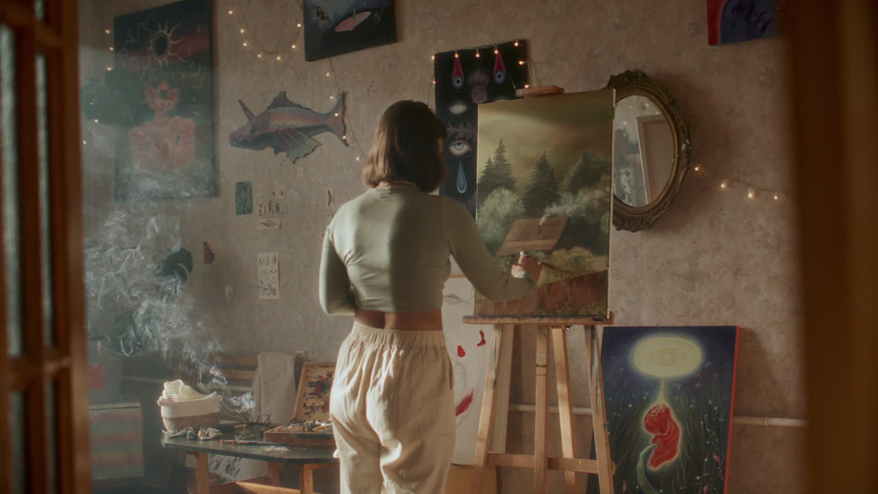 Ung, kvinnelig kunstner som maler landskapsscene på staffeli i et koselig rom