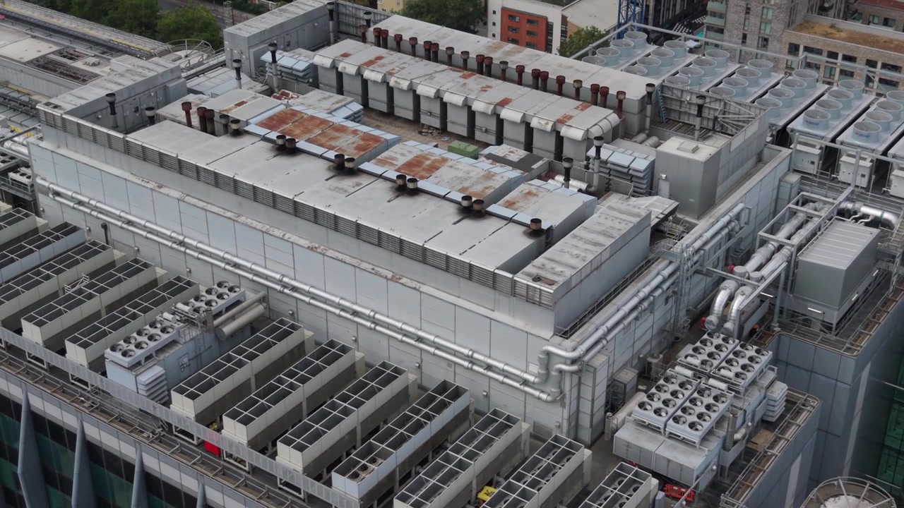 Luftfoto, der viser et stort moderne datacenter-tag. Talrige køleenheder og industrielt udstyr er spredt ud over anlægget i et bymiljø i lyse dagtimer.