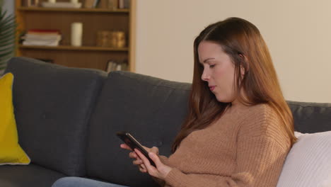 Woman-Sitting-On-Sofa-At-Home-Using-Mobile-Phone-To-Check-Social-Media-Message-And-Scrolling-Online-21