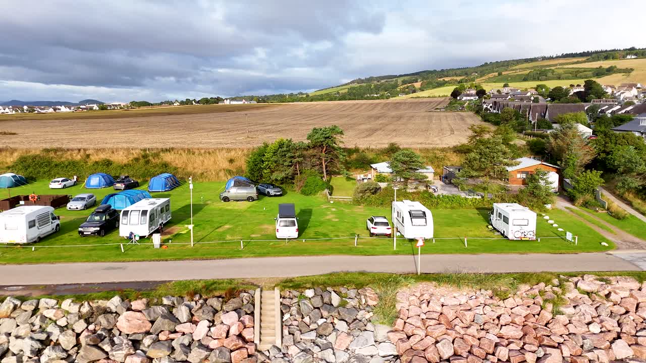 Drone footage pans across a coastal caravan and camping site with motorhomes, tents, and cars under partly cloudy daylight in Rosemarkie, Scotland