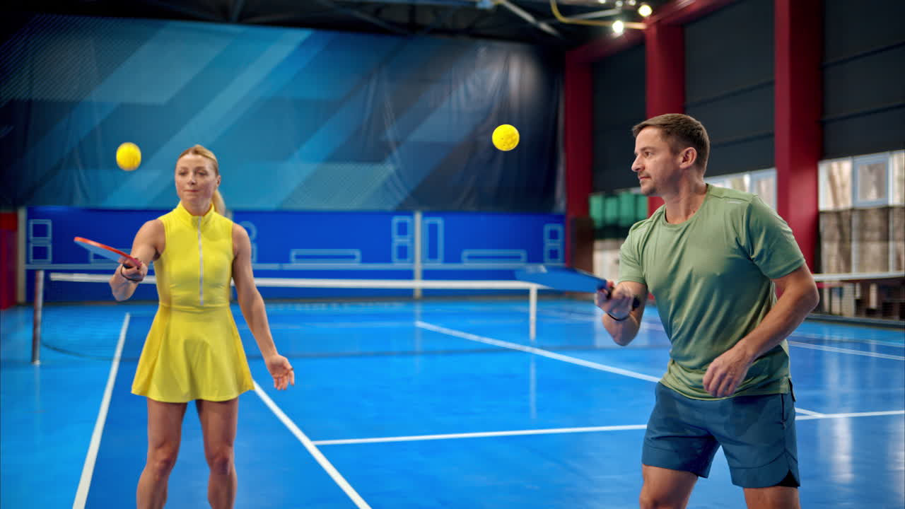A man and a woman training to play pickleball on a blue, inside court