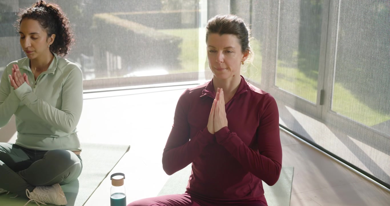 Diverse female friends doing yoga on mats by windows breathing then raising palms near teal flask