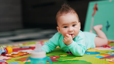 curious toddler in blue shirt lies on mat on his belly. adorable kid chews his thumb and looks around. blurred backdrop.