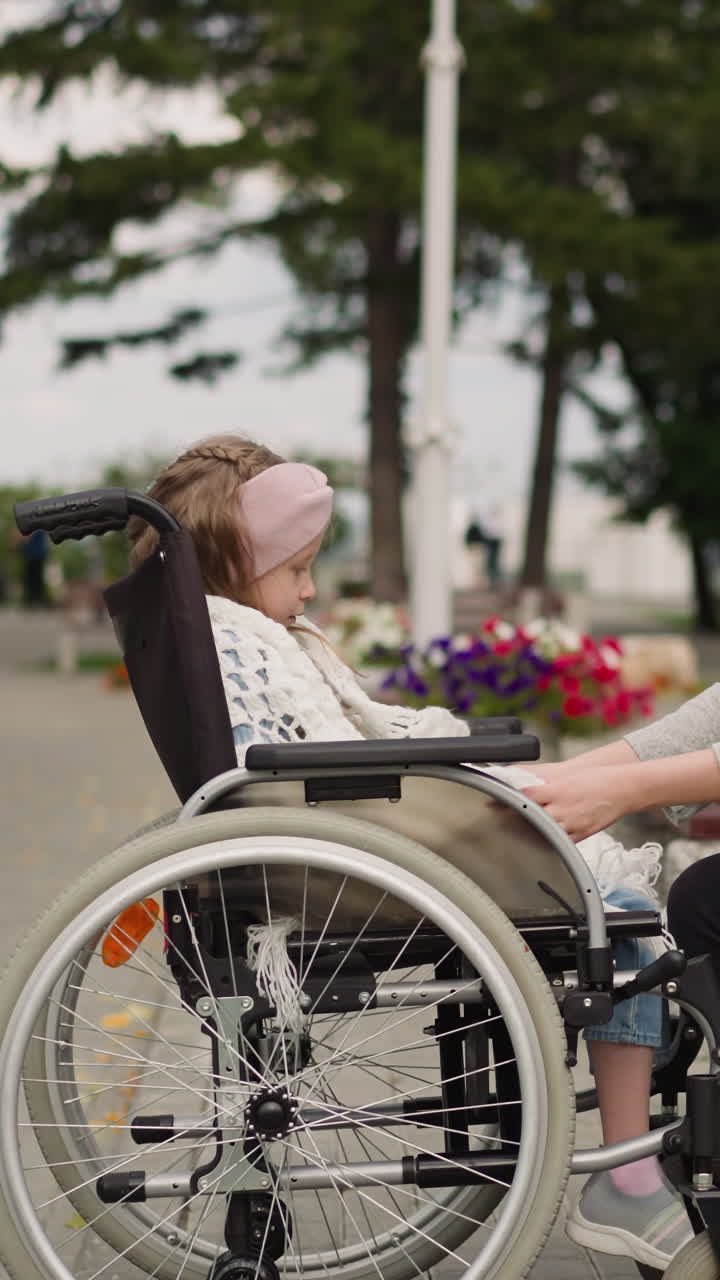 Caring mother wraps little daughter in warm knitted scarf sitting on bench in park. Young woman takes care of girl with injury in wheelchair