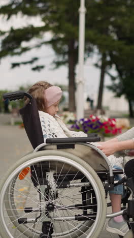 caring mother wraps little daughter in warm knitted scarf sitting on bench in park. young woman takes care of girl with injury in wheelchair