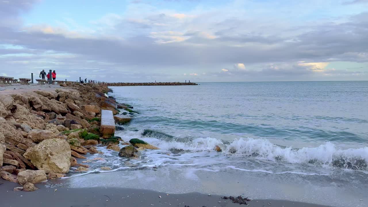 Rocky shoreline with gentle waves splashing against moss-covered rocks. A pier extends into the sea with people walking under a partly cloudy sky, capturing a serene coastal scene.