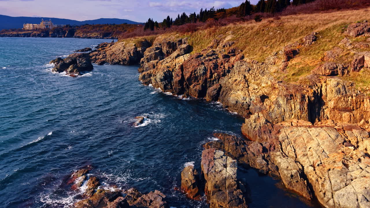 Scogliere costiere e onde lungo una costa frastagliata vicino a una città. Le onde si infrangono contro le rocce lungo la costa vicino a una piccola città durante le ore diurne.