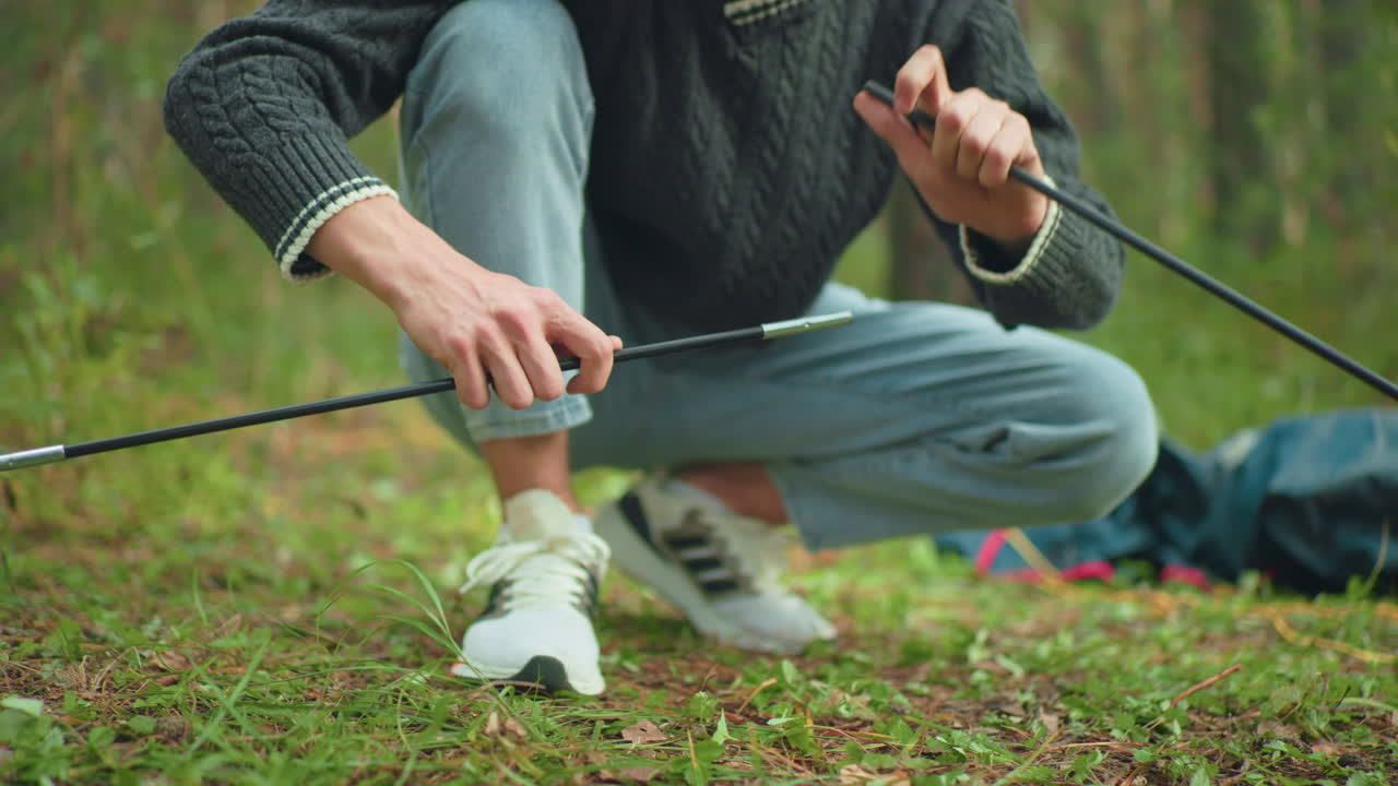 Close up of person handing tent poles to man in canvas squatting on forest floor as he joins parts together for camping setup with blurred background of foliage and scattered outdoor gear