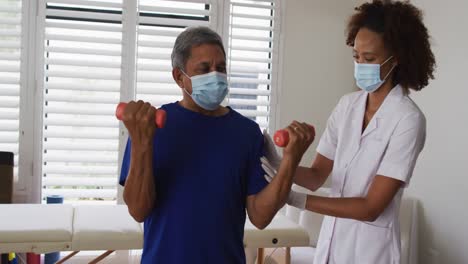 Mixed-race-female-physiotherapist-wearing-mask-helping-senior-exercise-using-dumbbells