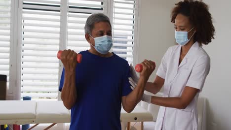 mixed race female physiotherapist wearing mask helping senior exercise using dumbbells