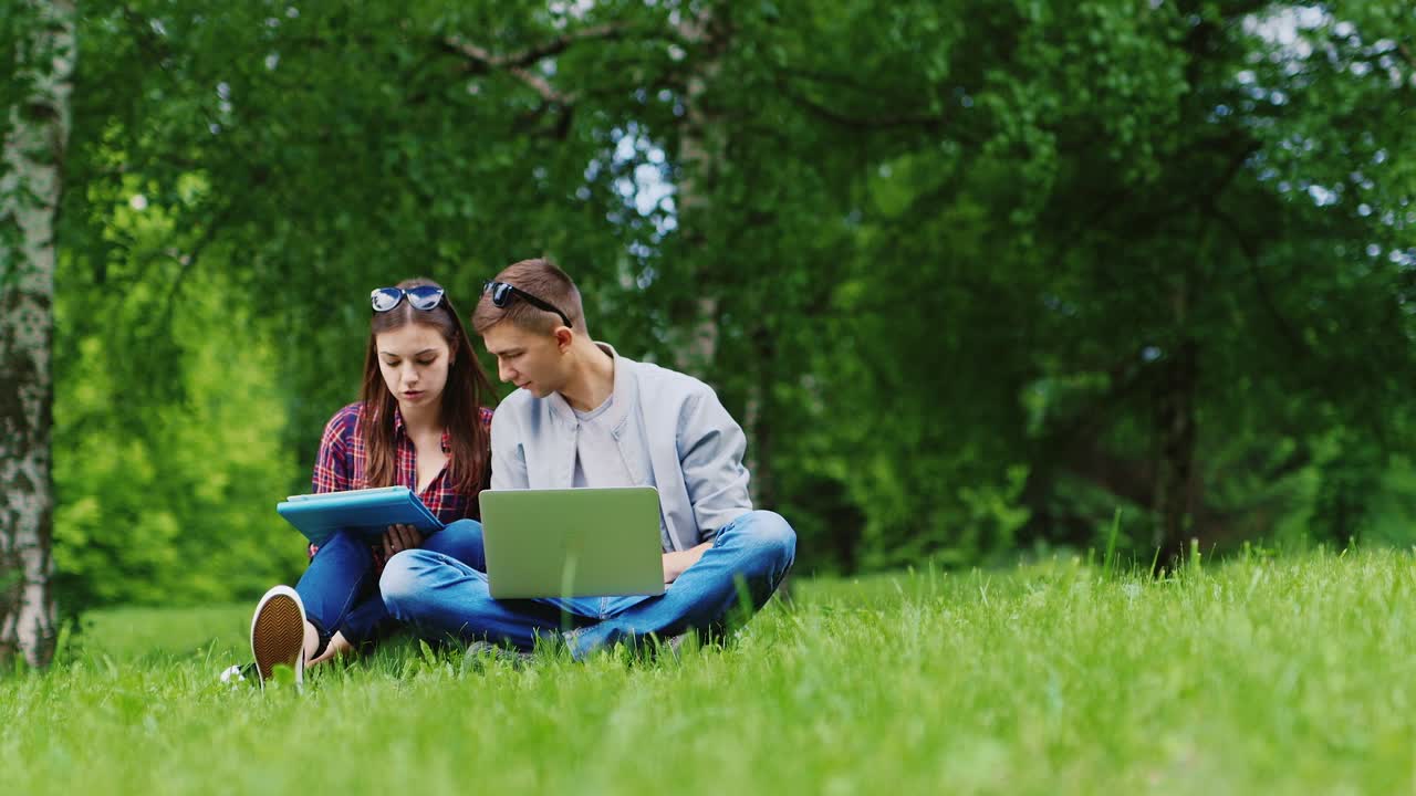 Ung mand og kvinde arbejder sammen på en bærbar computer og tablet, siddende på græsset i parken. HD-video