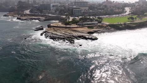 La-Jolla-coastline-landscape-over-ocean-in-California-with-crashing-tide-wave-on-jagged-rock,-parallax-aerial-near-San-Diego