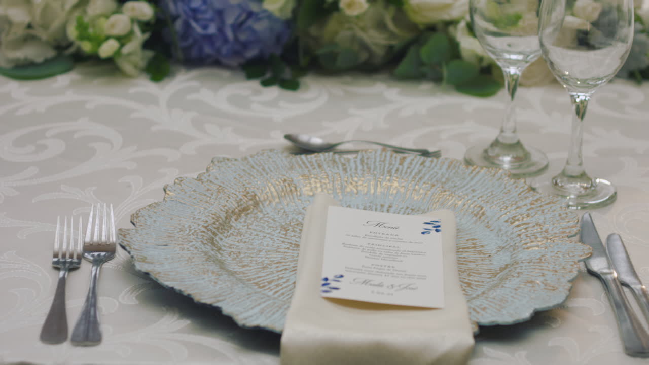A detailed clip of an elegantly decorated wedding table. It features stemware, a white tablecloth, plates, cutlery, cloth napkins, a delicate floral arrangement, and a personalized invitation.